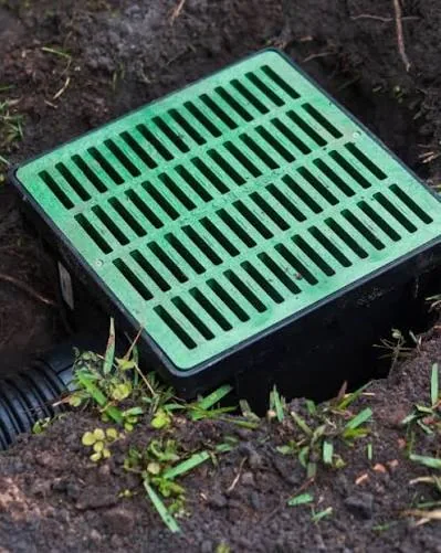 Square catch basin with a green grate set level with the soil, connected to a corrugated drain pipe