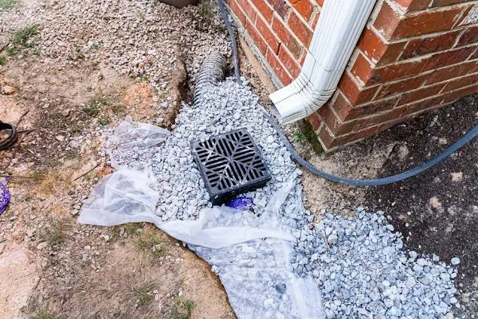 Catch basin set in washed gravel on filter fabric beside a downspout, with the drain pipe running away from the house