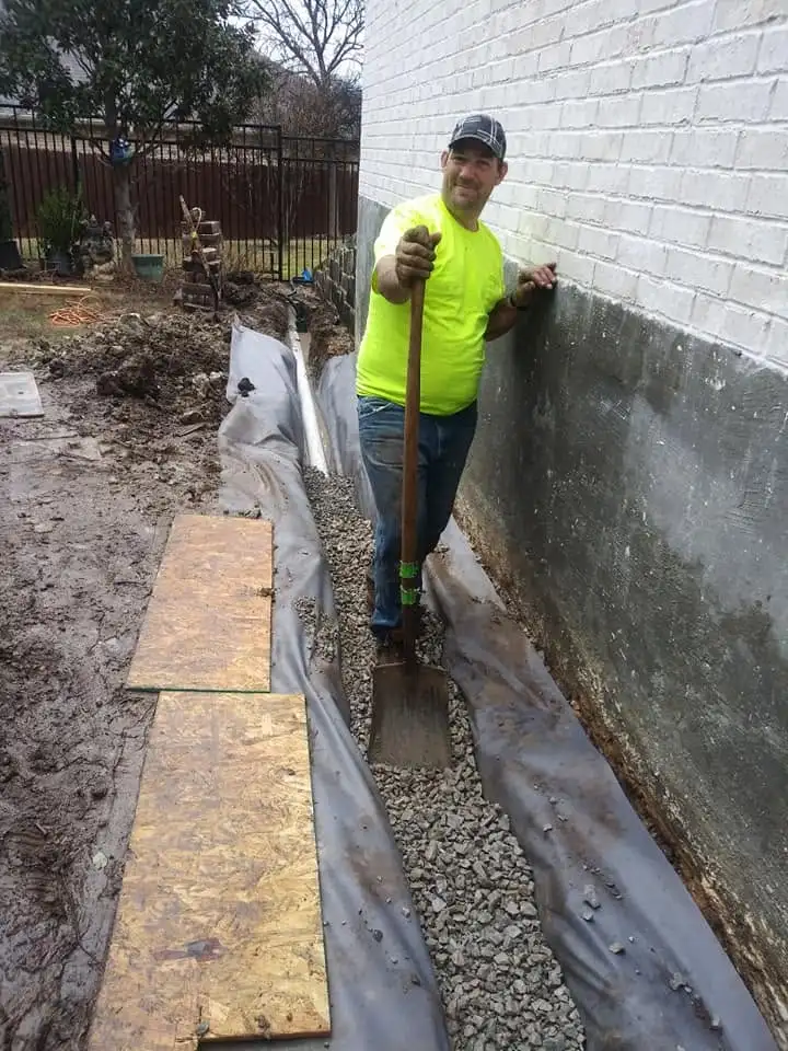Lone Star Irrigation technician digging a French drain trench lined with filter fabric beside a house