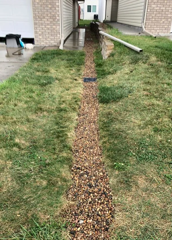 Finished gravel-topped drain line running down a side yard between two homes, with a catch basin grate at the downspout
