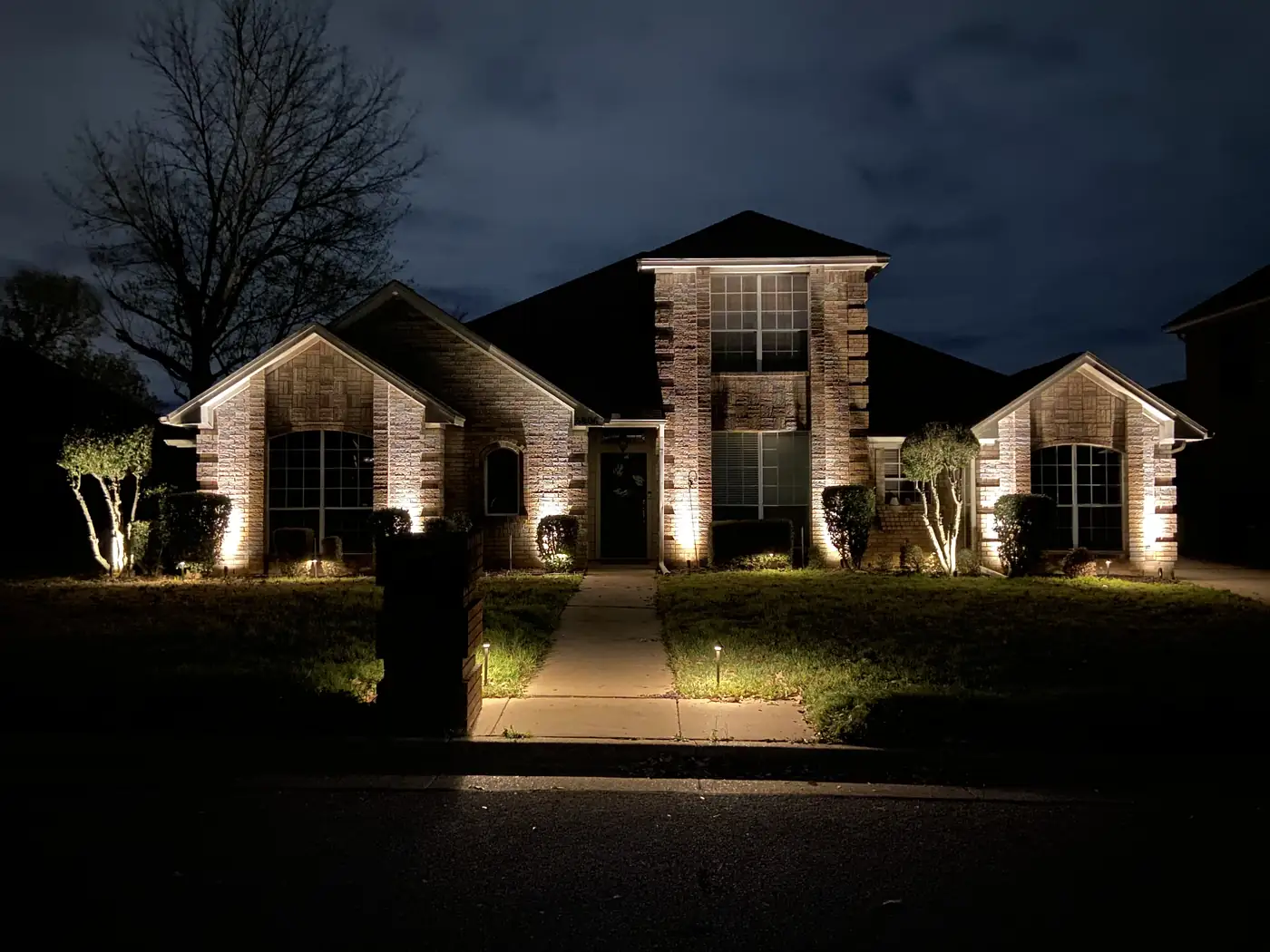 Front of a brick home at night with uplit trees and lights along the beds