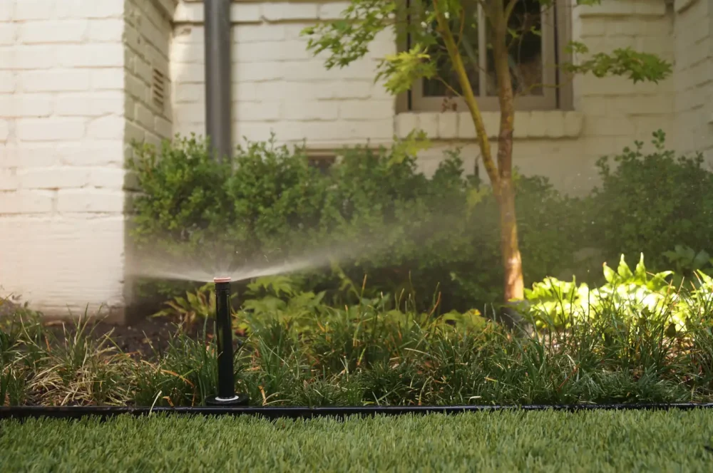 Sprinkler head watering a garden bed beside a painted brick home